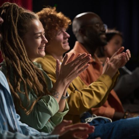 Group of diverse people sitting in row and applauding during a performance, capturing joy and engagement, showcasing various ethnic backgrounds and unity