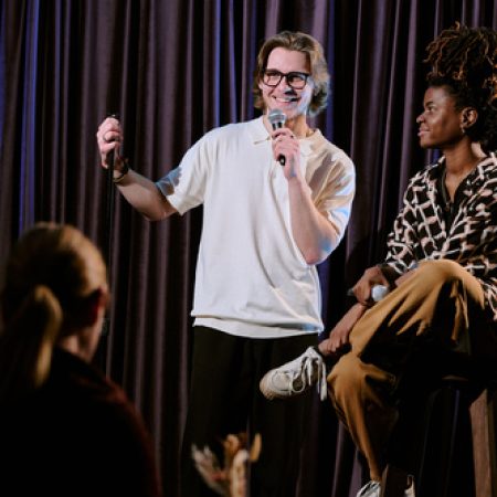 Comedian performing stand-up comedy routine on stage holding microphone while another person sitting nearby observing performance at comedy club