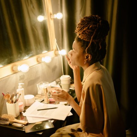 Woman preparing for performance in dressing room illuminated by stage lights, surrounded by makeup tools and notes. Reflective moment before stepping on stage