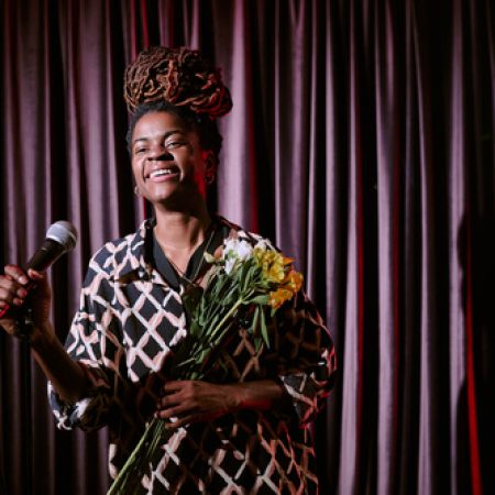 African American performer smiling while holding microphone and bouquet of flowers on stage during performance with long dreadlocks and patterned outfit