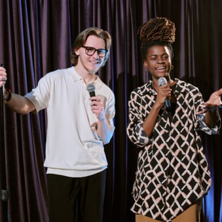 Young man and woman holding microphones, smiling warmly while performing comedy in front of a dark curtain, engaging audience with humor and charisma